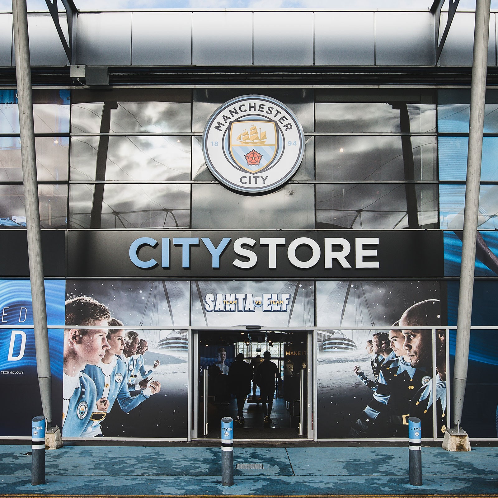 A photograph of the front of the Manchester City Stadium shop. On the front window a version of the main campaign image showing, Team Santa stood opposite Team Elf, in the middle the doorway to the shop