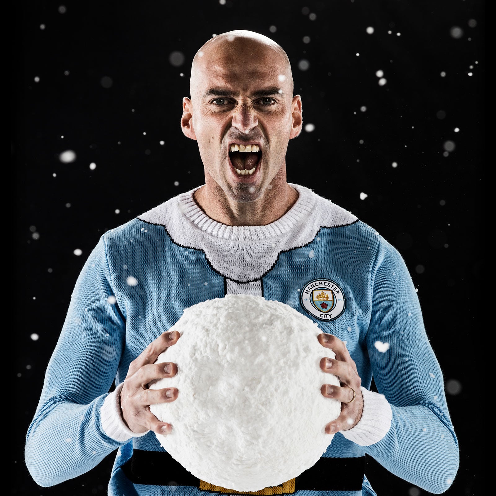 Will Caballero stood close to camera, holding a giant snowball shouting towards camera. Snow is falling over a black background behind him